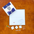 A top-down view of a board game set labeled 'BUILD GAME BOX' on a glossy orange burl wood surface. The set includes a white box with blue panels, a white game board, a small red die, a printed instruction sheet, several white square tiles, and small white player pieces.