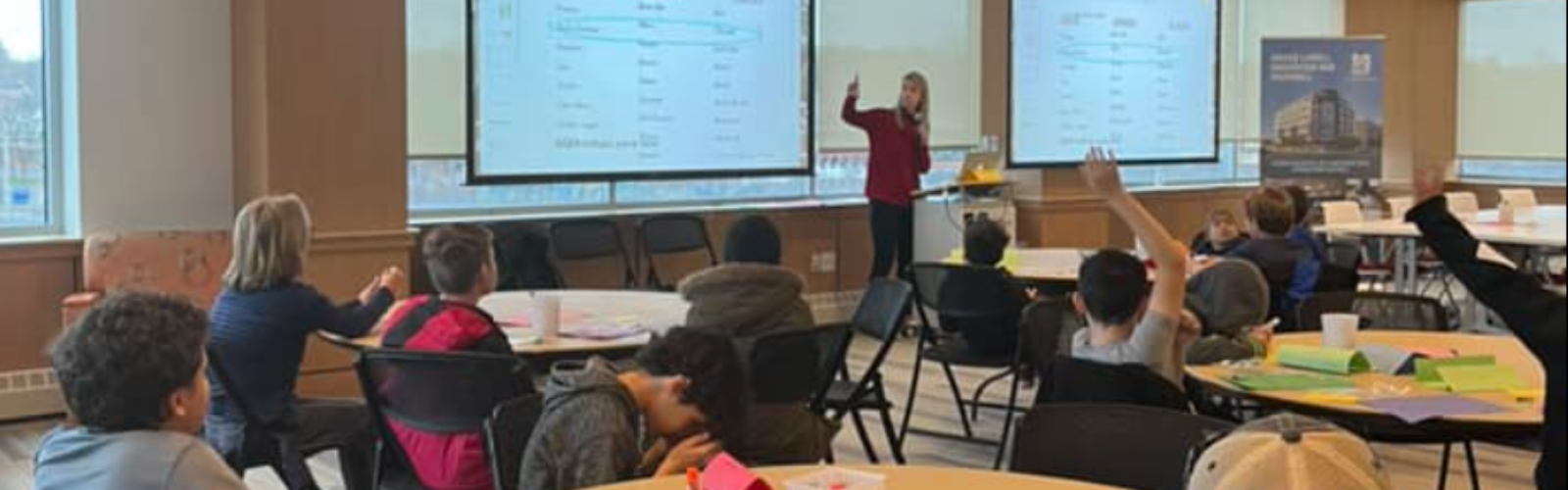 A game design classroom scene with a presenter standing in front of two large projection screens, with children and adults seated at tables and raising hands.