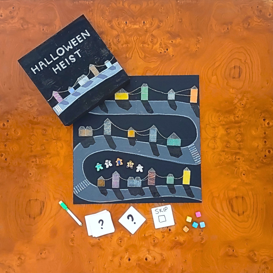 A flat-lay view of a board game set on a glossy orange burl wood surface. The set includes a white and blue box labeled 'BUILD GAME BOX', a white game board, a small red dice, white tiles, player tokens, and a green marker.