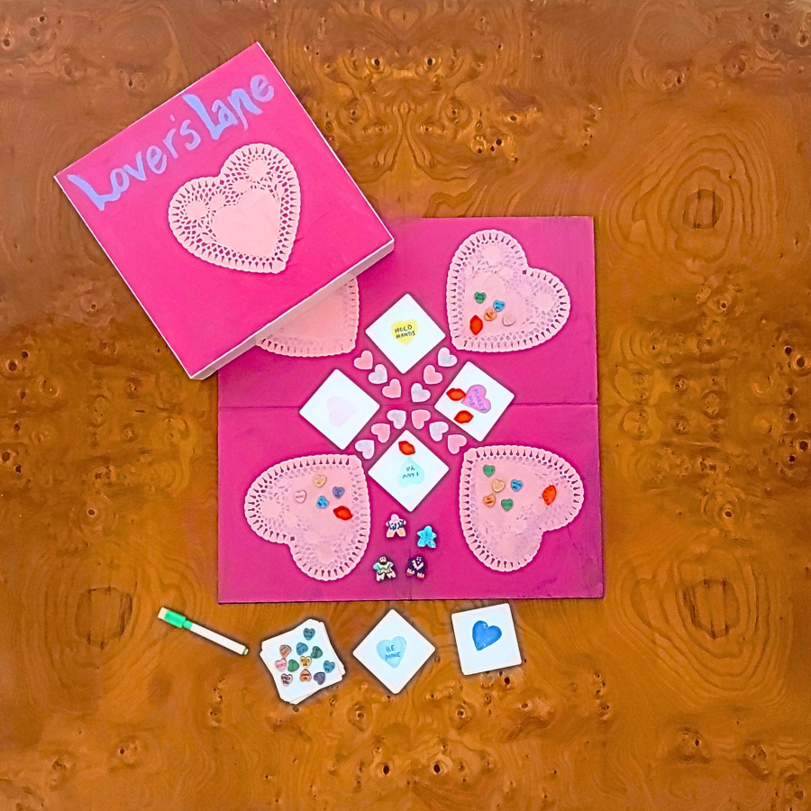 A pink heart-themed craft or game set laid out on a textured brown surface, with a matching pink box labeled 'Love's Lane' placed above it. The set includes heart-shaped fabric pieces, small white square cards, decorative embellishments, and a small green marker.
