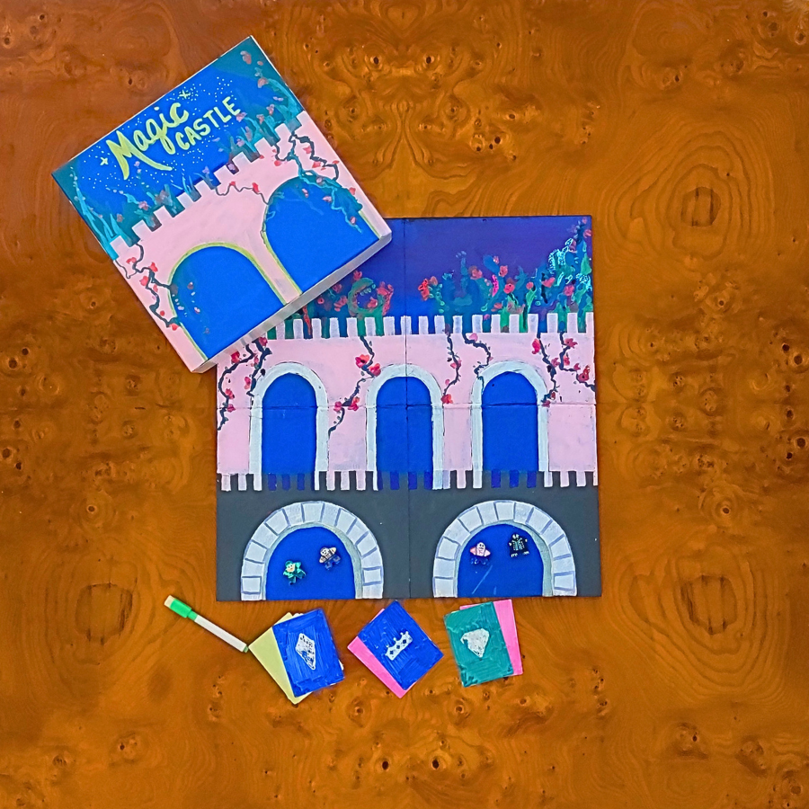 A Magic Castle toy set arranged on a textured orange-brown wood surface, including a colorful box, a partially assembled castle board, and several small accessory cards or tiles.