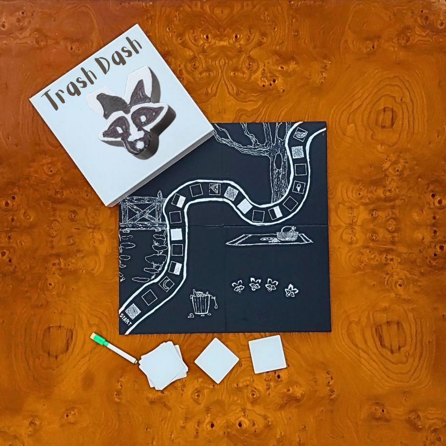 Trash Dash board game set laid flat on a textured orange wood background, including a white game box with a raccoon logo, a dark play mat with white illustrated path and icons, three white cards, and a small green-and-white marker.